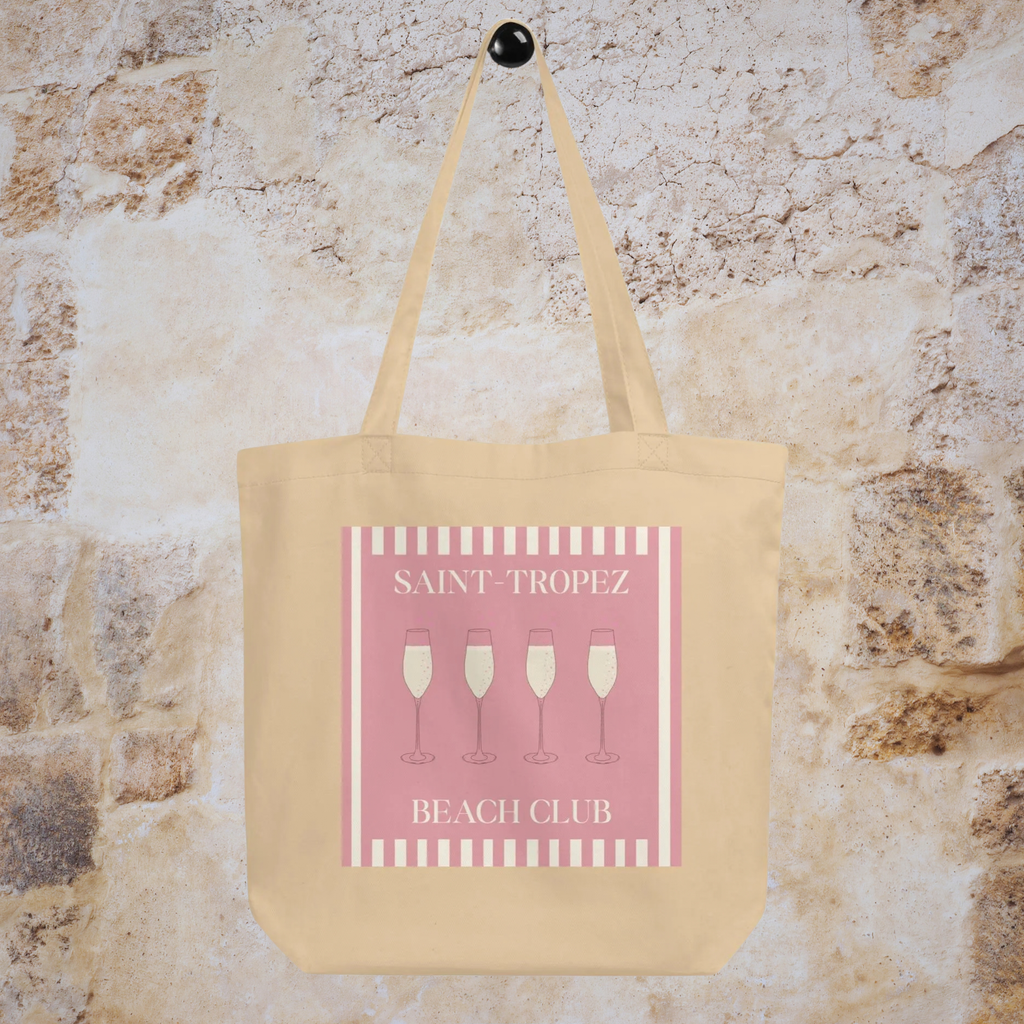 Saint-Tropez Beach Club tote hanging against a pale stone wall, showing the full pink graphic.