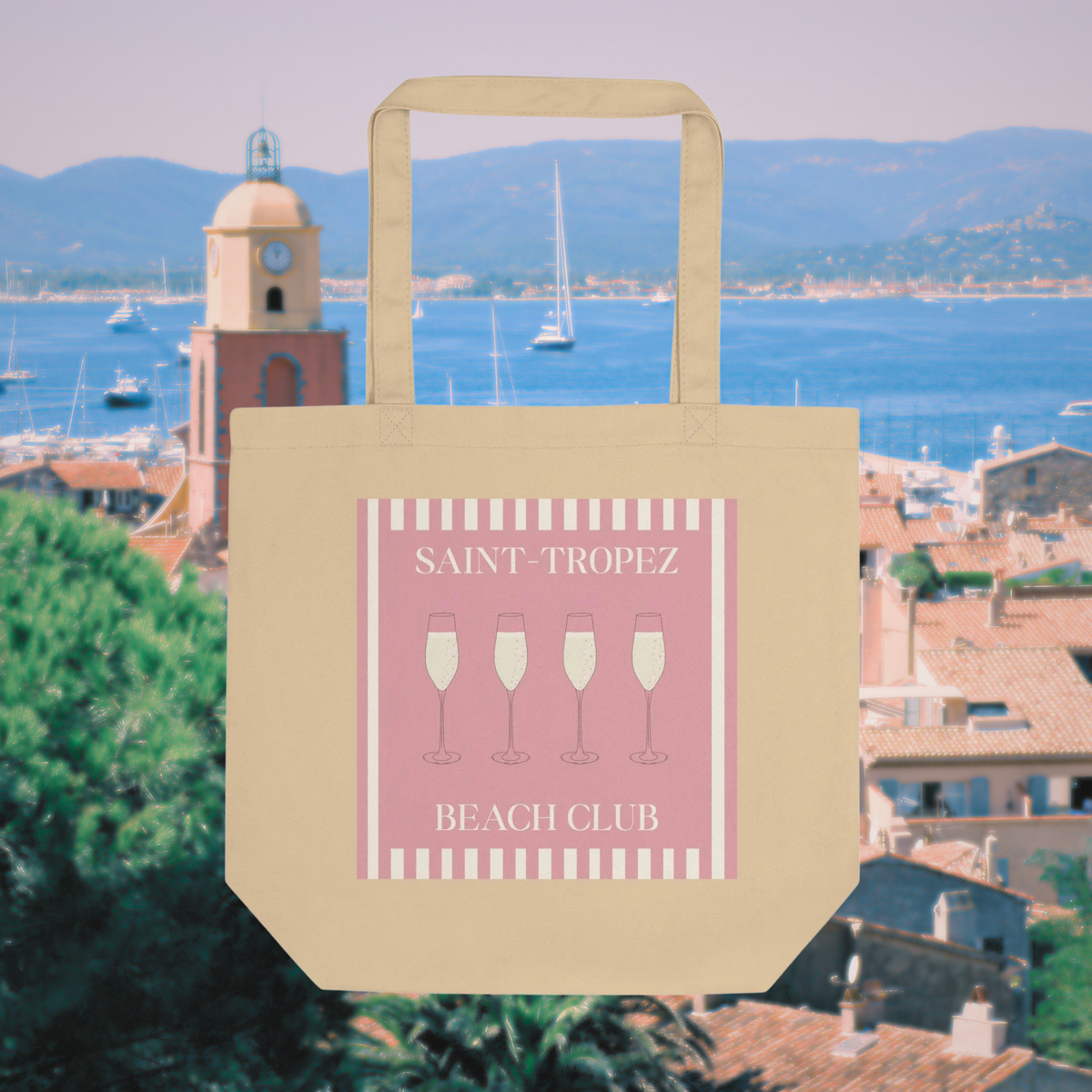 Natural cotton Saint-Tropez Beach Club tote with a pink striped print and champagne glasses, shown above the harbour.