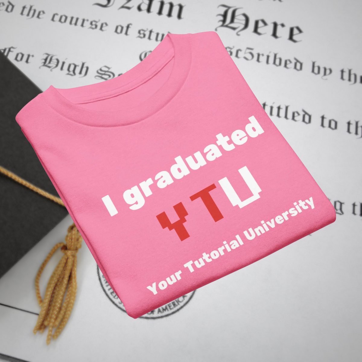 I Graduated YTU kids' T-shirt in pink, folded beside a diploma and graduation cap.