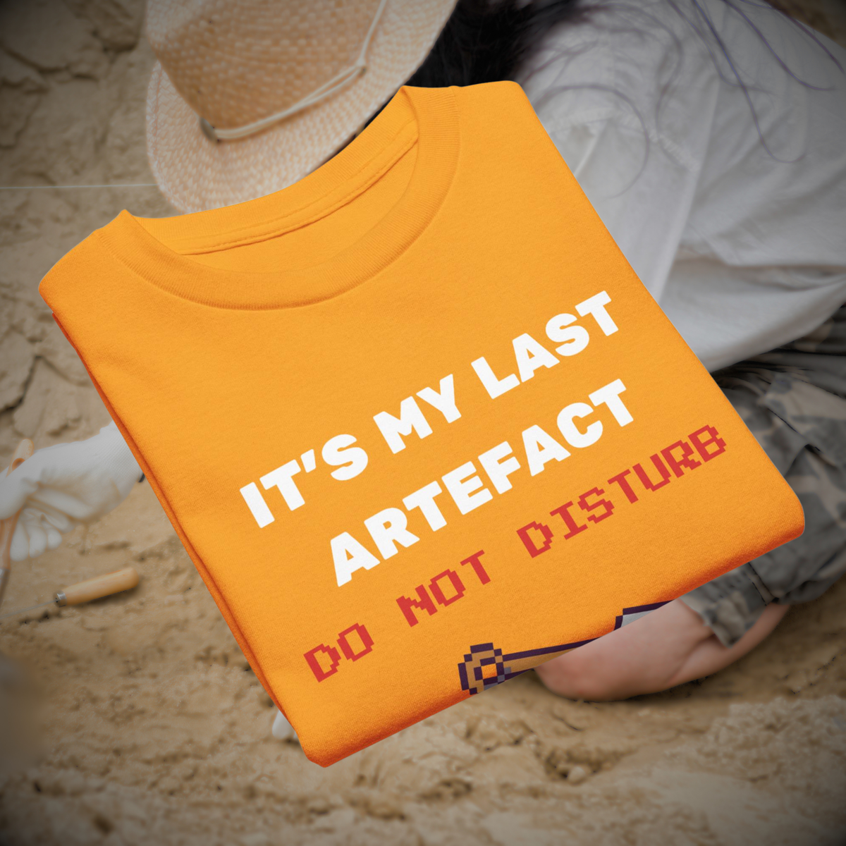 It's My Last Artefact kids' gamer T-shirt in gold, folded beside archaeological tools and a hat.