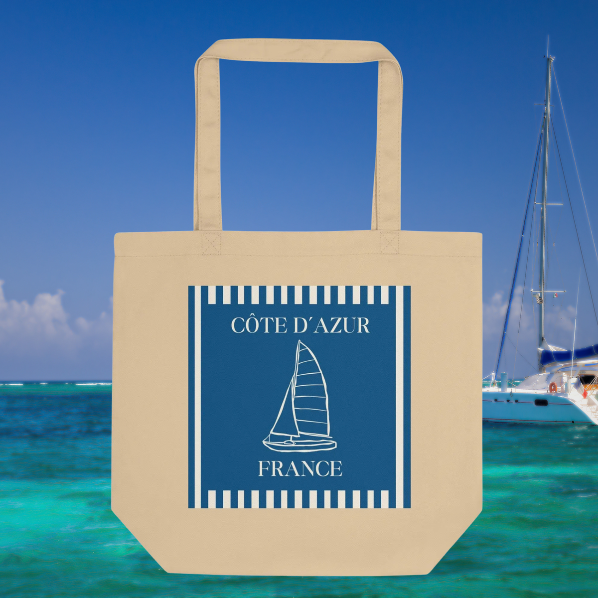 Natural cotton Côte d’Azur tote with a blue striped panel and white sailboat graphic beside a marina.