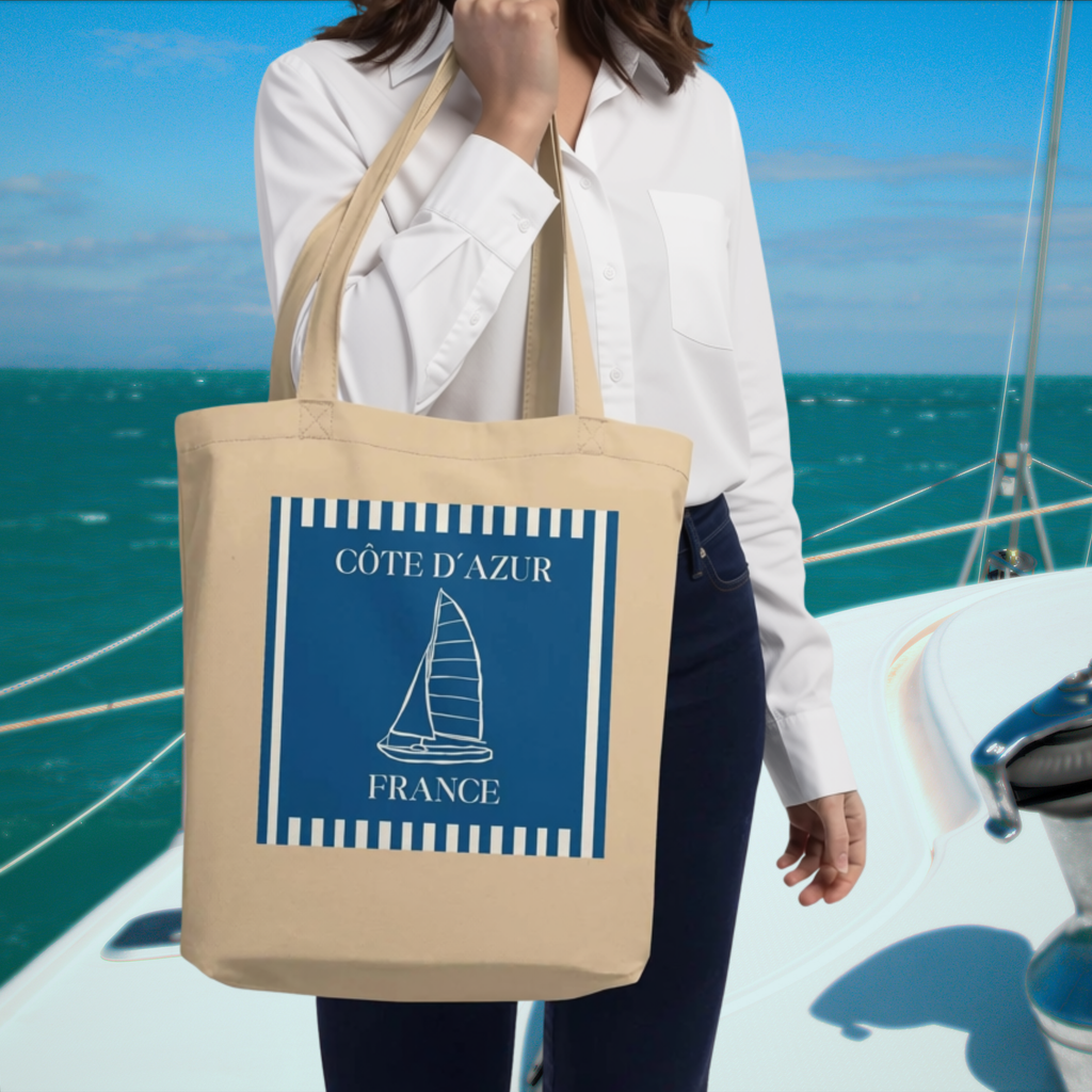 Woman carrying the blue Côte d’Azur sailboat tote on a boat beside turquoise water.