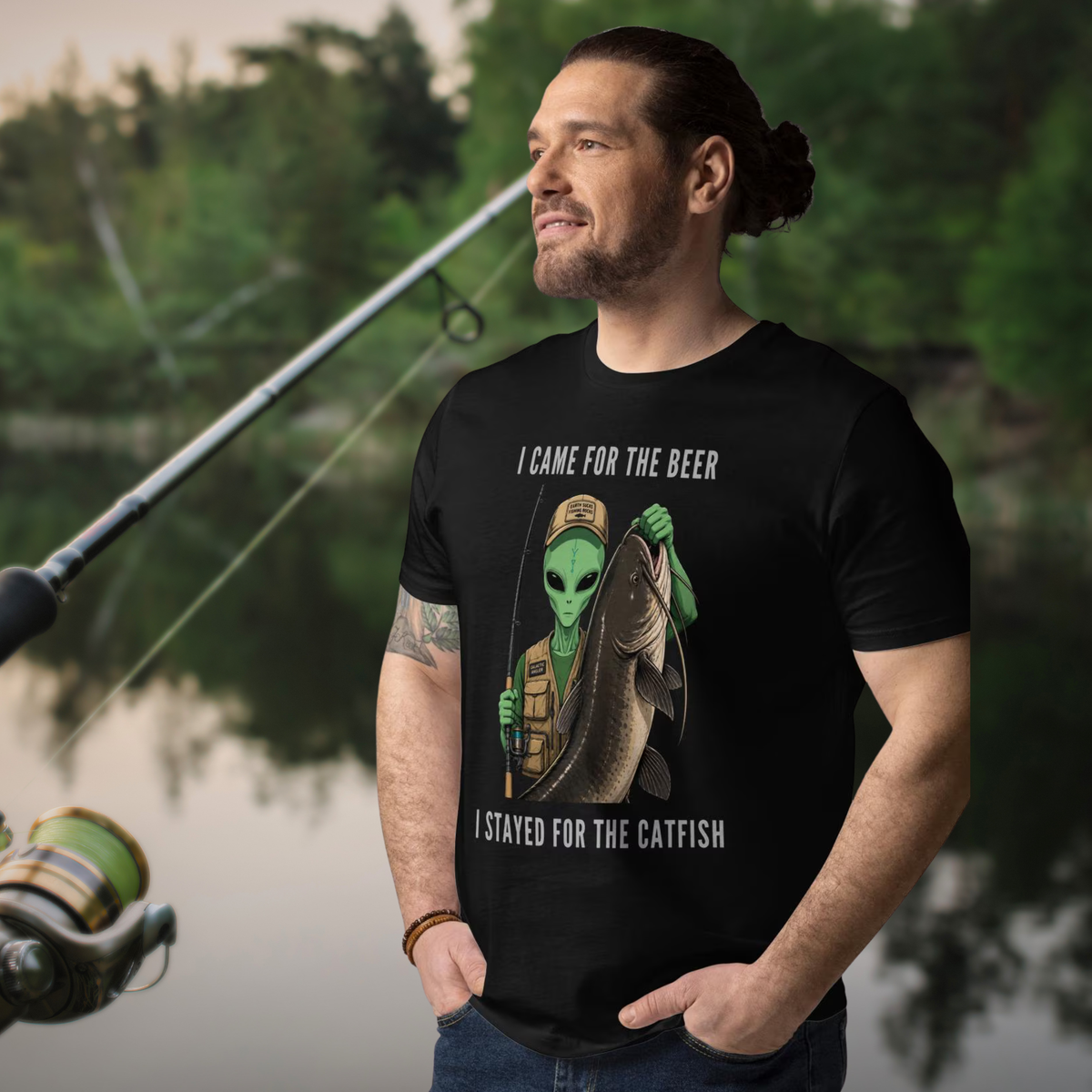 Alien Catfish fishing T-shirt in black, worn by a model beside a fishing rod and water.