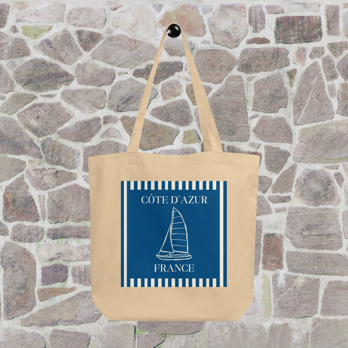Côte d’Azur sailboat tote hanging against a stone wall, showing the full blue and white graphic.